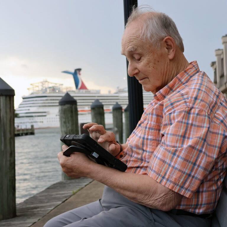 An elderly man in a plaid shirt sits on a dock by the water, using a tablet, with a large cruise ship docked in the background.