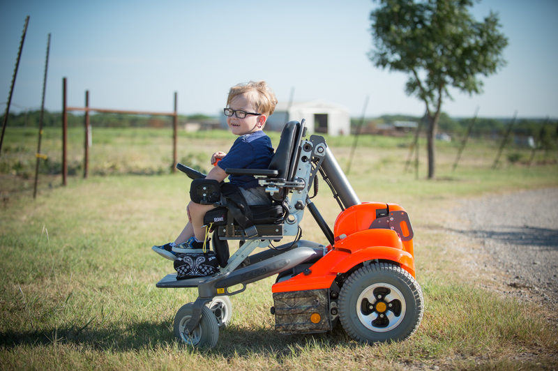 A young child with glasses sits in an orange pediatric power wheelchair on a grassy path outdoors, with a fence, tree, and open field in the background on a sunny day.