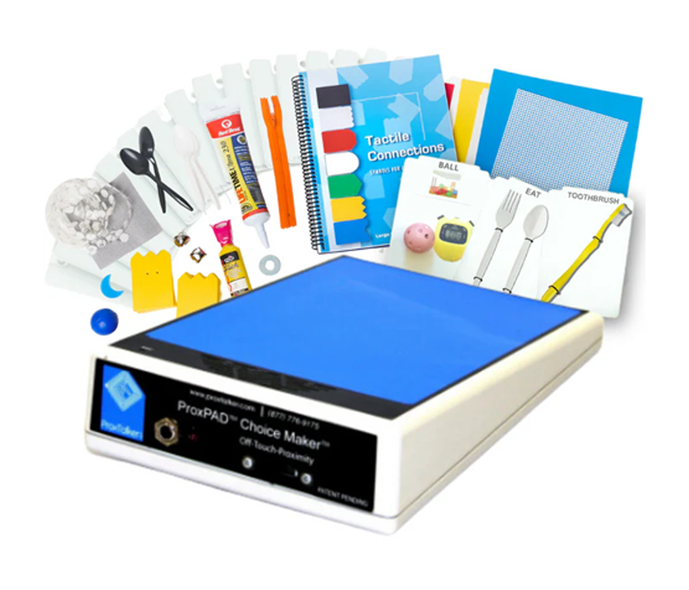An assistive communication device with a blue touchpad sits among educational materials, including cards, a notebook, scissors, craft supplies, and instructional booklets designed for accessible learning.