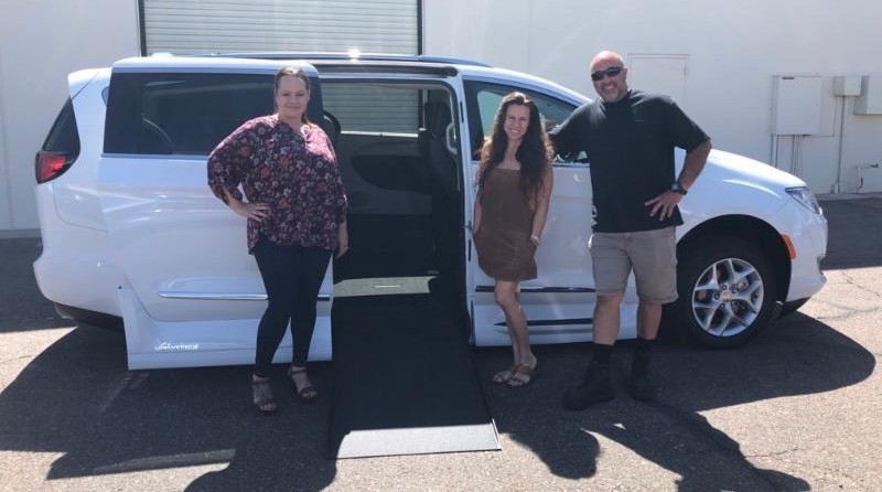 Three people stand next to a white wheelchair accessible vehicle with a side ramp extended. The woman on the left wears a floral top, the woman in the middle a brown dress, and the man on the right sunglasses and shorts—all smiling at the camera.