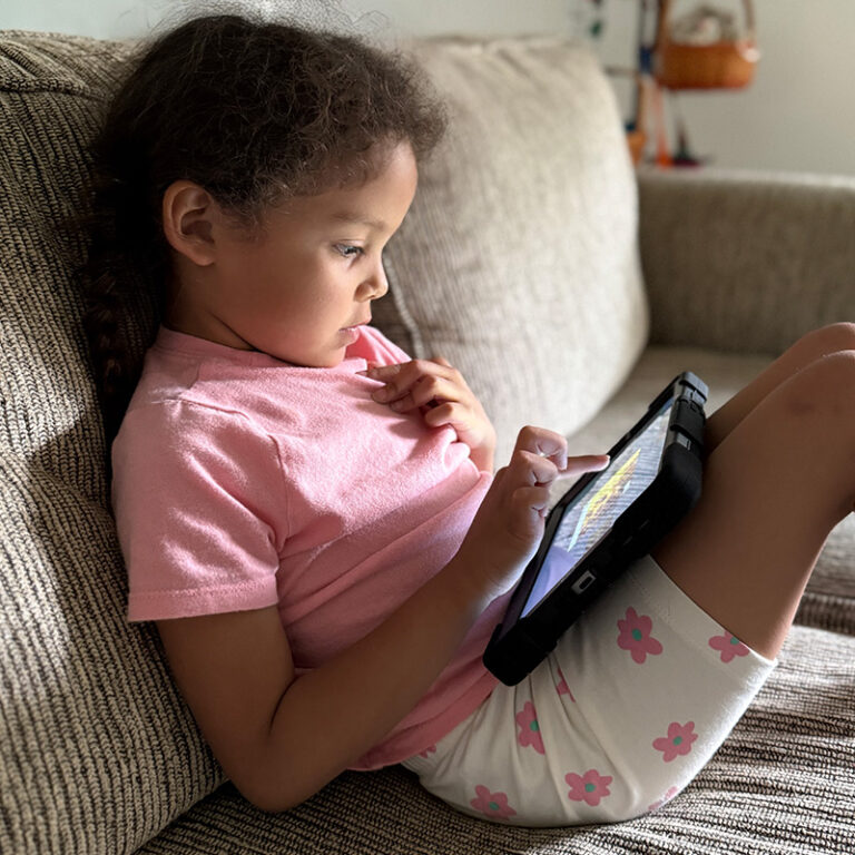 A young girl with curly hair sits on a couch, wearing a pink shirt and flower-print shorts, intently exploring a tablet with her finger.