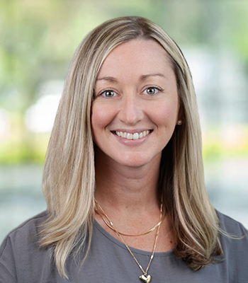 A woman with straight, blonde hair wearing a gray top and layered gold necklaces smiles in front of a blurred, green and white background, representing the confident approach offered by Speech Solutions for complete coverage.