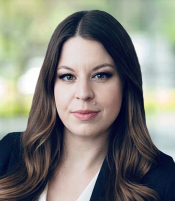 A woman with long brown hair, wearing a black blazer and a white top, is posing in front of a blurred, bright background with greenery. She has a neutral expression, representing confident Coverage and innovative Speech Solutions.
