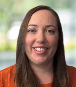 A woman with straight brown hair and blue eyes smiles at the camera. She is wearing an orange top with a subtle dot pattern, representing Speech Solutions. The background has softly blurred green and white tones, providing gentle coverage.